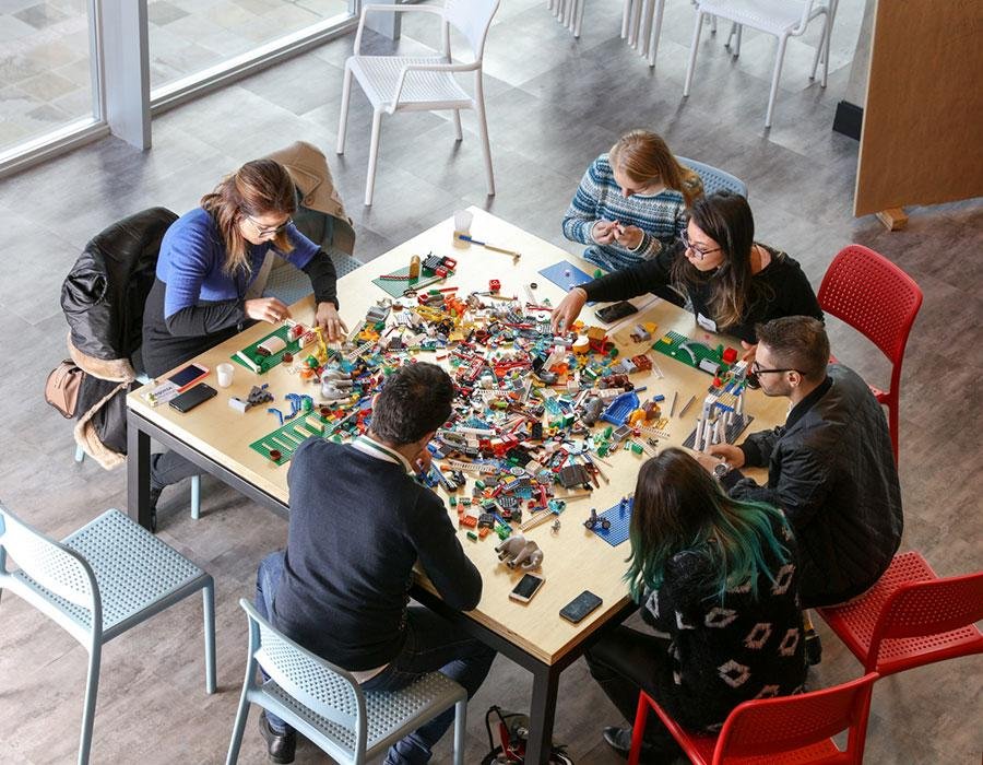 Personas sentadas a una mesa cuadrada con piezas de lego esparcidas.